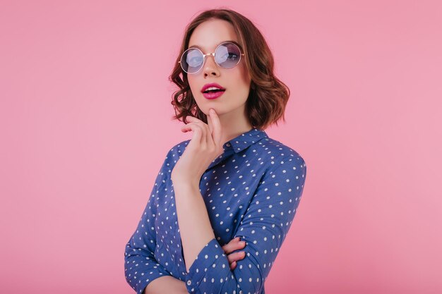 Chica caucásica refinada con gafas de sol moradas posando con expresión de cara interesada Mujer joven jocunda con cabello oscuro escalofriante durante la sesión de fotos sobre fondo rosa