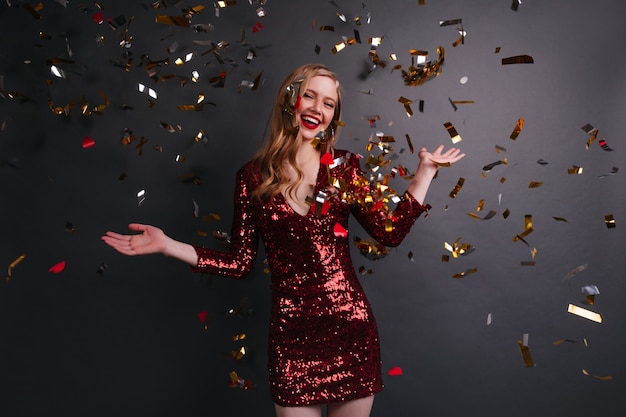 Chica caucásica refinada en vestido rojo bailando en la fiesta. Disparo de estudio de linda mujer rubia posando bajo confeti.