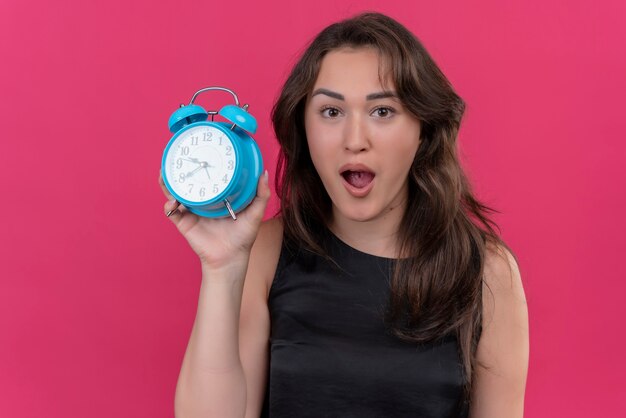 Chica caucásica sorprendida vistiendo camiseta negra sosteniendo un reloj despertador sobre fondo rosa