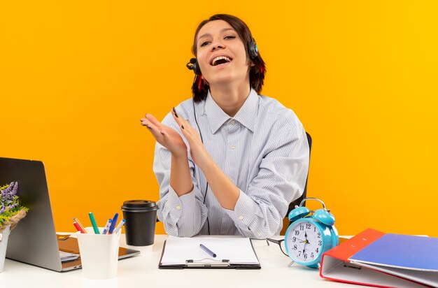 Chica de centro de llamadas joven alegre con auriculares sentado en el escritorio aplaudiendo aislado en naranja