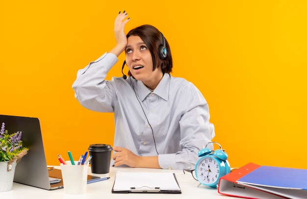 Chica de centro de llamadas joven confundido con auriculares sentado en el escritorio poniendo la mano en la frente mirando hacia arriba aislado en naranja