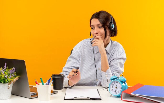 Chica de centro de llamadas joven pensativa con auriculares sentado en el escritorio con herramientas de trabajo poniendo el dedo en los labios mirando portátil aislado en la pared naranja