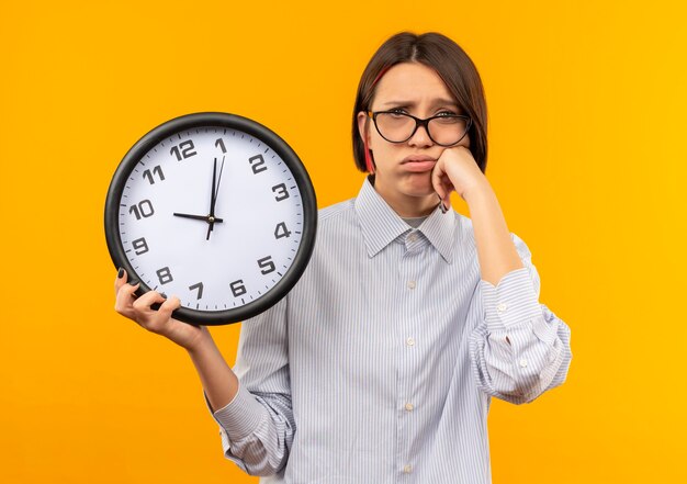 Chica de centro de llamadas joven triste con gafas sosteniendo el reloj poniendo la mano en la mejilla aislada en la pared naranja