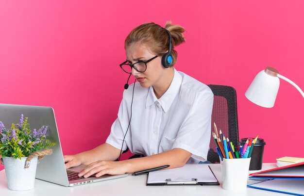 Chica de centro de llamadas rubia joven concentrada con auriculares y gafas sentado en el escritorio con herramientas de trabajo usando una computadora portátil aislada en la pared rosa