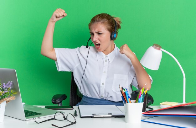 Chica de centro de llamadas rubia joven confiada con auriculares sentado en el escritorio con herramientas de trabajo mirando portátil haciendo gesto fuerte aislado en la pared verde