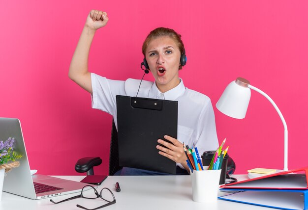 Chica de centro de llamadas rubia joven confiada con auriculares sentado en el escritorio con herramientas de trabajo sosteniendo el portapapeles mirando a la cámara haciendo gesto de sí aislado en la pared rosa