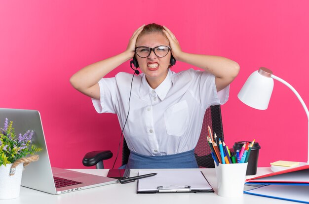 Chica de centro de llamadas rubia joven irritada con auriculares y gafas sentado en el escritorio con herramientas de trabajo manteniendo las manos en la cabeza mirando a cámara mostrando los dientes aislados en la pared rosa