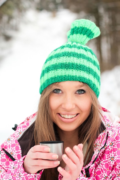 Chica en una chaqueta carmesí y sombrero verde beber té caliente de un termo en las montañas cubiertas de nieve