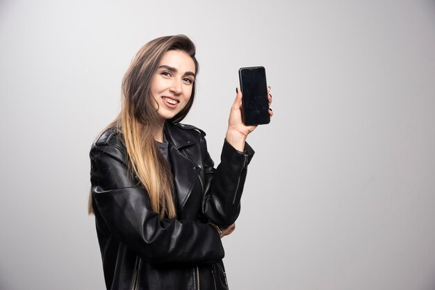 Chica con chaqueta de cuero sosteniendo un teléfono inteligente sobre un fondo gris.