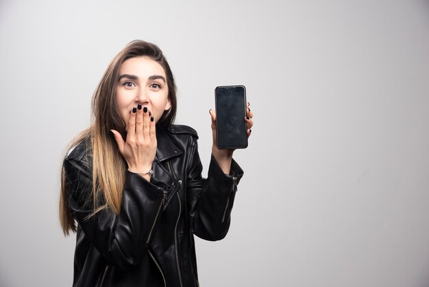 Chica con chaqueta de cuero sosteniendo un teléfono inteligente sobre un fondo gris.