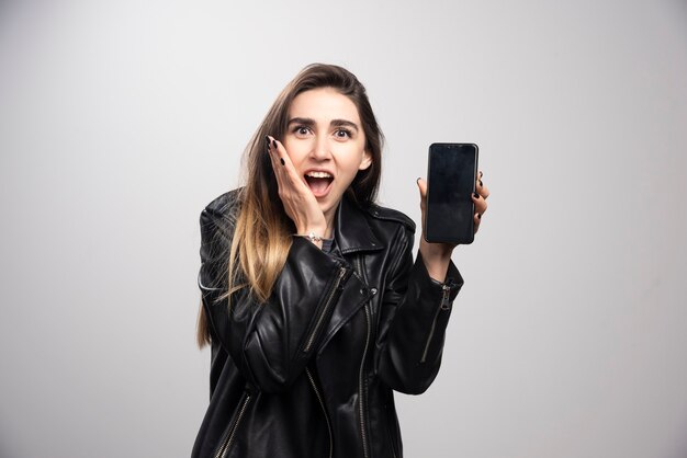 Chica con chaqueta de cuero sosteniendo un teléfono inteligente sobre un fondo gris.