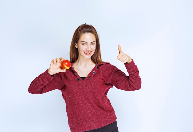 Chica de chaqueta roja sosteniendo una manzana roja y mostrando un signo de mano positivo