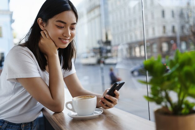 Chica charlando en el teléfono inteligente y tomando café en el restaurante cerca de la ventana, sonriendo a la pantalla del teléfono celular