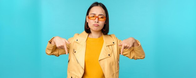 Una chica china triste y sombría con gafas de sol señala con el dedo hacia abajo y hace una mueca se ve miserable y decepcionada por la emoción de arrepentimiento de pie sobre un fondo azul