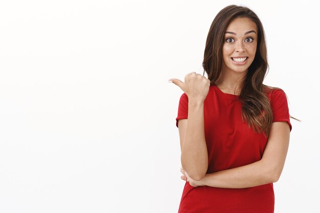 Chica chismeando sobre algo extraño que vio detrás, de pie emocionada y entusiasta con una camiseta roja, apuntando con el pulgar a la izquierda espacio de copia en blanco en blanco, sonriendo intrigada, de pie en la pared del estudio