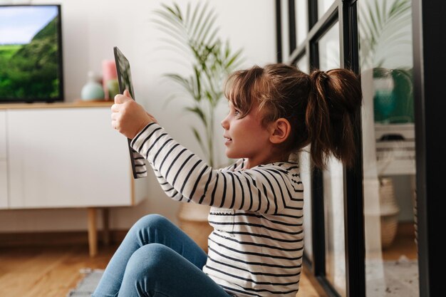 Chica con cola de caballo se toma selfie en la sala de estar y sostiene una tableta