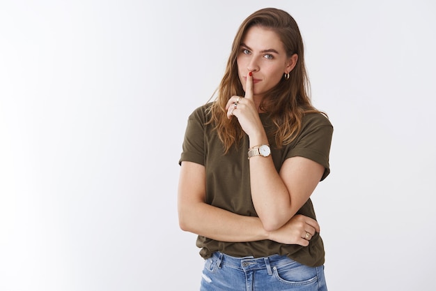 Chica compartiendo secretos de belleza, prometo no contarle a nadie. Encantador modelo femenino europeo lindo tortuoso mostrando silencio pidiendo silencio de pie fondo blanco dedo índice presionó la boca, sonriendo