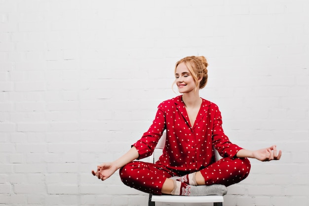 Chica complacida en pijama rojo haciendo yoga en la pared blanca. Retrato interior de una joven rubia sentada en posición de loto en una silla.