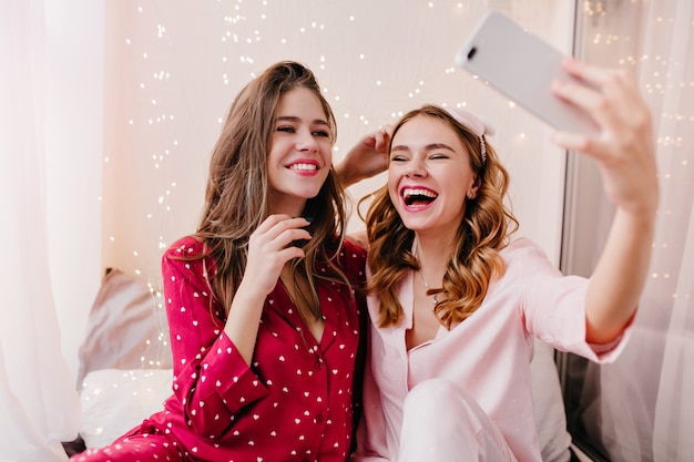 Chica complacida en pijama rosa divirtiéndose en su habitación con su mejor amiga. Señora joven alegre haciendo selfie en la mañana con su hermana.