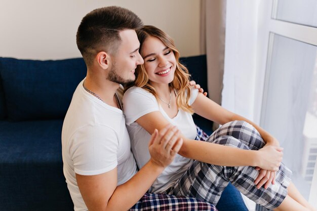 Chica complacida en ropa de dormir linda sentada en el sofá con su novio y sonriendo. Filmación en interiores de modelo femenino de ensueño pasando el fin de semana con su marido.