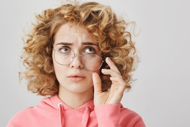 Chica complicada y desconcertada con gafas dobladas mira la esquina superior derecha pensativa