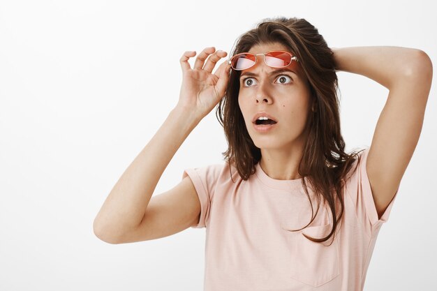 Chica confundida con gafas de sol posando contra la pared blanca