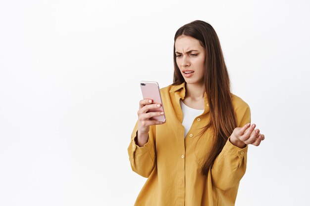 Chica confundida mirando la pantalla del teléfono y haciendo muecas de disgusto, mensaje extraño en el chat, no puedo entender algo, de pie sobre una pared blanca