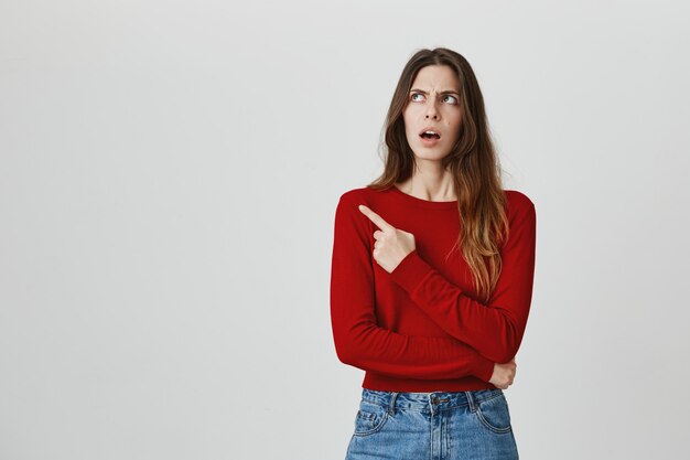 Chica confundida y perpleja, frunciendo el ceño perplejo, señalando y mirando la esquina superior izquierda