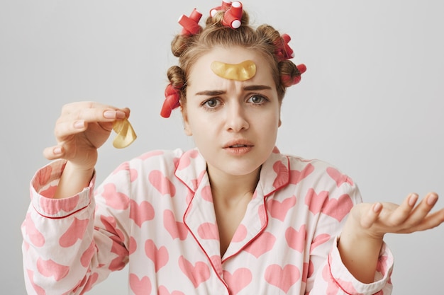 Chica confundida en rizadores de pelo y pijama tratando de aplicar parches en los ojos