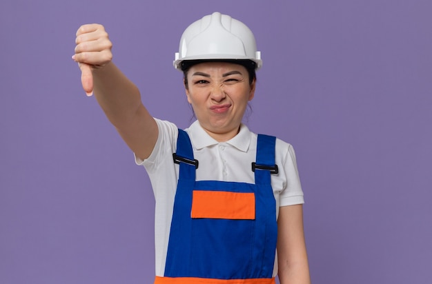 Chica constructora asiática joven disgustada con casco de seguridad blanco apuntando hacia abajo