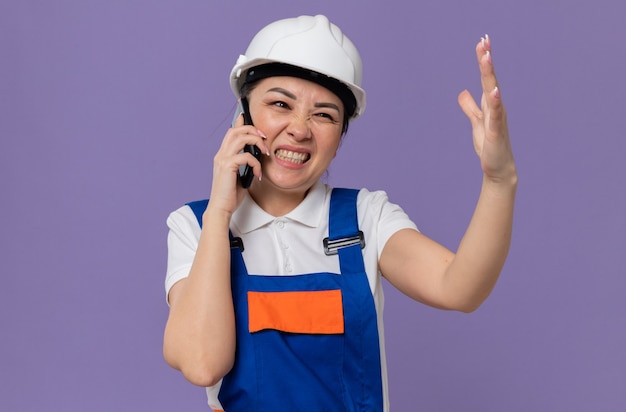 Chica constructora asiática joven molesta con casco de seguridad blanco gritando a alguien en el teléfono manteniendo la mano abierta