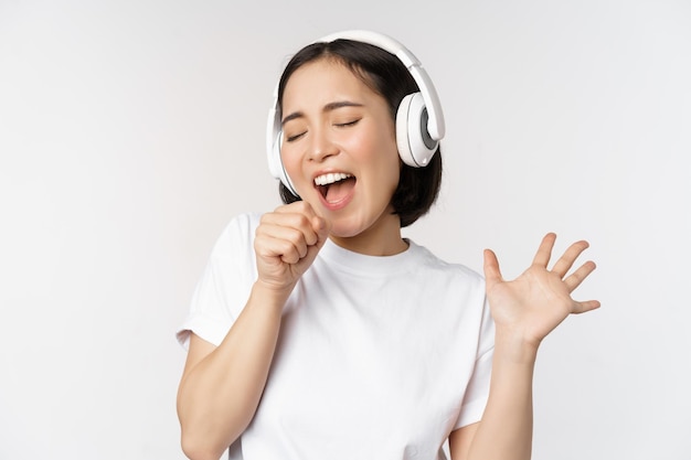 Chica coreana canta y escucha música en auriculares divirtiéndose sobre fondo blanco.