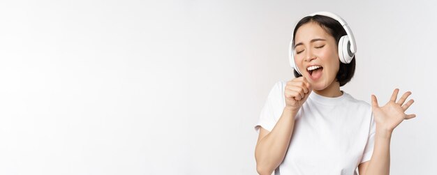 Chica coreana canta y escucha música en auriculares divirtiéndose sobre fondo blanco.