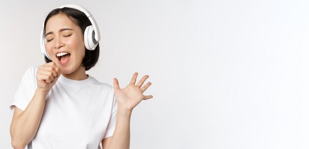 Chica coreana canta y escucha música en auriculares divirtiéndose sobre fondo blanco.