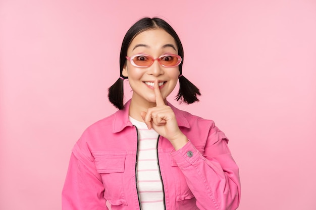 Chica coreana moderna con elegantes gafas de sol de traje de primavera que muestran el signo de callar, presionar el dedo en los labios, gesto tabú de pie sobre fondo rosa