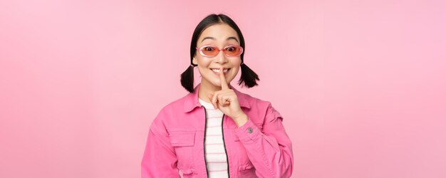 Chica coreana moderna con elegantes gafas de sol de traje de primavera que muestran el signo de callar, presionar el dedo en los labios, gesto tabú de pie sobre fondo rosa