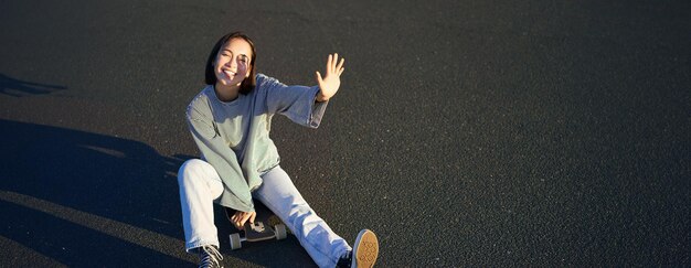 Foto gratuita la chica coreana positiva se cubre la cara de la luz del sol se sienta en el patineta y sonríe felizmente