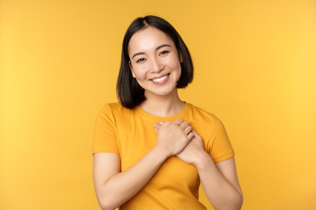 Chica coreana romántica mujer asiática cogida de la mano en el corazón sonriendo con cuidado y ternura de pie sobre fondo amarillo