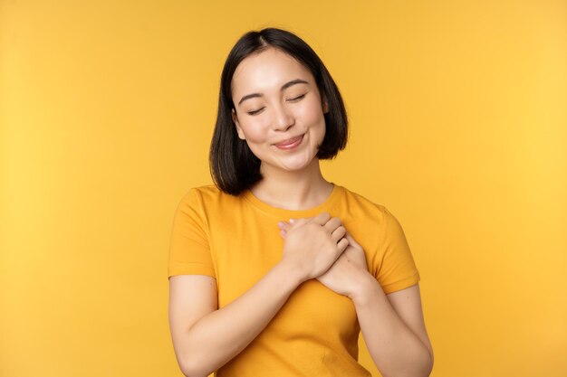 Chica coreana romántica mujer asiática cogida de la mano en el corazón sonriendo con cuidado y ternura de pie sobre fondo amarillo