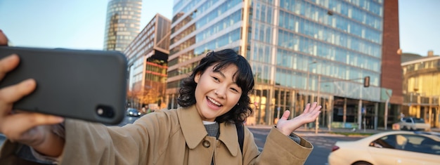 Chica coreana sonriente mostrando la ciudad tomando selfie frente a los edificios en la calle con cara feliz