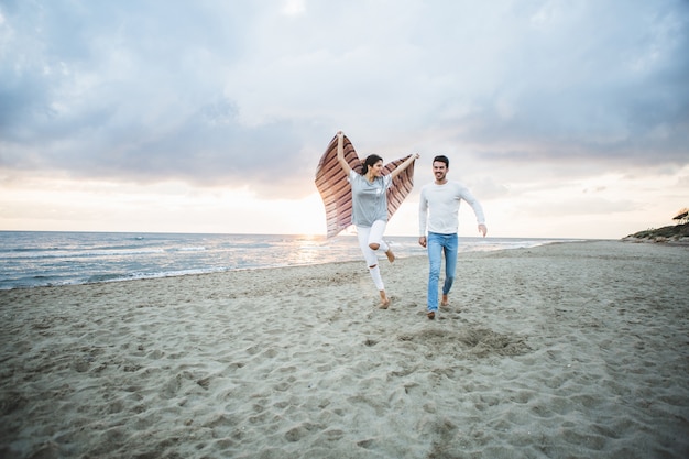 Chica corriendo por la playa con una manta y su novio al lado