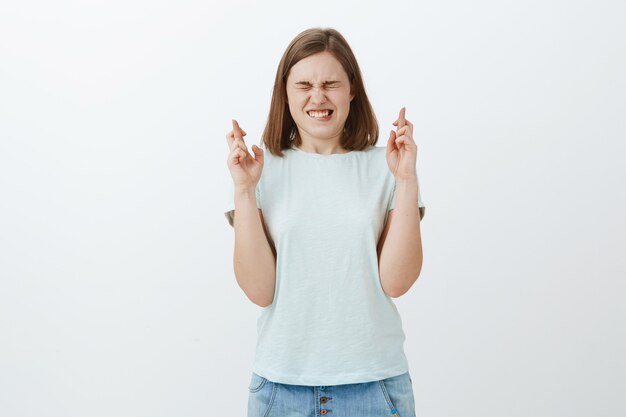 Chica cree en el milagro rezando y pidiendo deseos cruzando los dedos para la buena suerte mientras cierra los ojos y aprieta los dientes poniendo todo su esfuerzo en soñar hecho realidad de pie intensamente sobre la pared gris