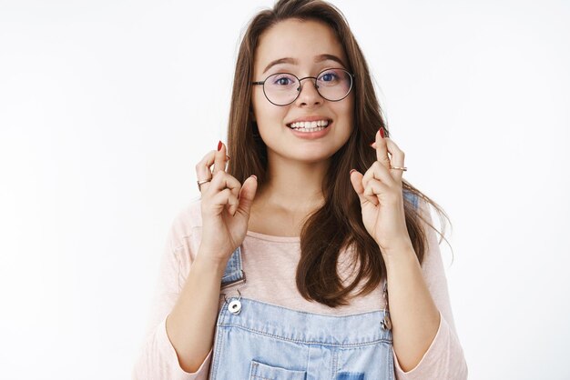 Chica cruzando los dedos con una sonrisa optimista y esperanzada que tiene un sueño de fe hecho realidad deseando lograr objetivos de pie fiel con expresión feliz emocionada en gafas y overoles de mezclilla