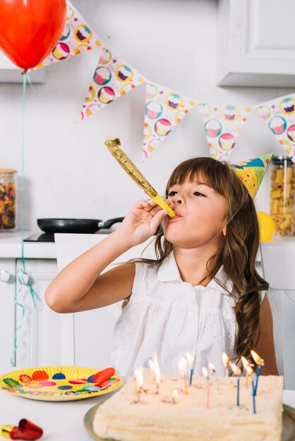 Chica de cumpleaños que sopla partido cuerno sentado detrás del pastel de cumpleaños con velas