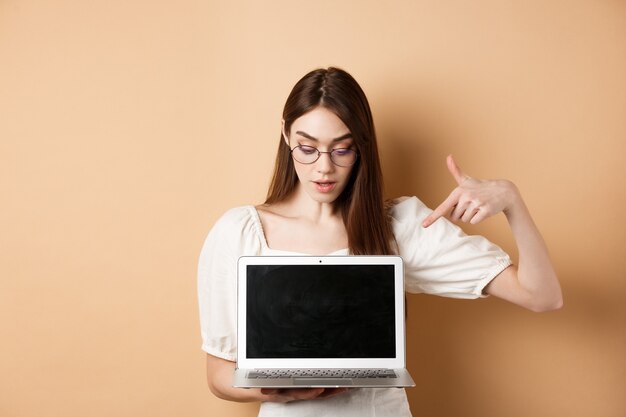 Chica curiosa con gafas apuntando a la pantalla de la computadora portátil echa un vistazo a la oferta en línea demuestre el proyecto en la computadora ...