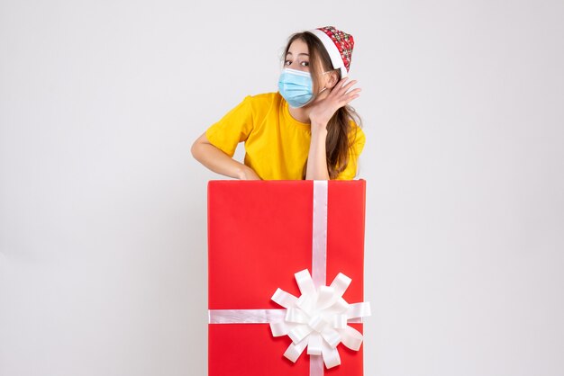 Chica curiosa con gorro de Papá Noel escuchando algo parado detrás de un gran regalo de Navidad en blanco