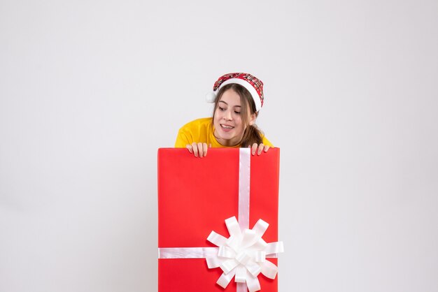 Chica curiosa con gorro de Papá Noel mirando algo de pie detrás de un gran regalo de Navidad en blanco