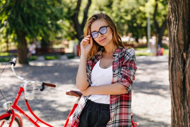 Chica curiosa en traje de primavera de pie junto a la bicicleta. Señora caucásica atractiva que presenta con la bicicleta en el parque.