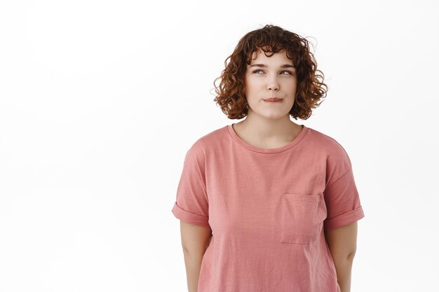 Chica curvilínea pensativa con cabello rizado, pensando, mirando a un lado mientras reflexiona, tomando la decisión de qué pedir la entrega de alimentos en línea, lamiendo los labios, de pie sobre fondo blanco.