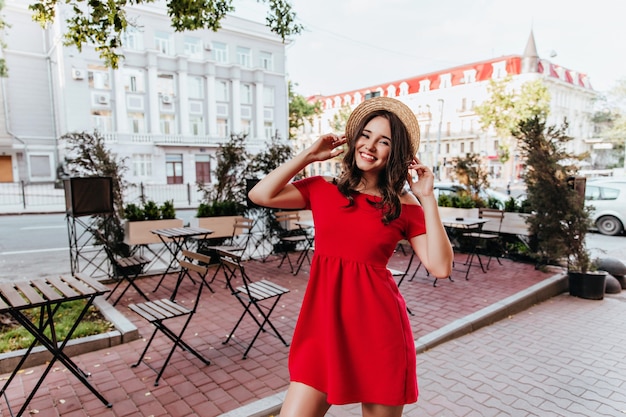 Chica delgada alegre posando junto a la cafetería de la calle. Emocionada mujer caucásica con sombrero de paja riendo durante la sesión de fotos de la ciudad.